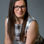 Katie is a White woman sitting facing the camera in front of a grey backdrop. She is wearing a white dress with music print, has long dark hair over her shoulders, and black square glasses.